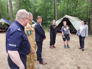 Zdjęcie w kolorze, przedstawia grupę osób w tym funkcjonariuszy policji i straży pożarnej stojących w grupie na obszarze leśnym, w tle namiot harcerski.