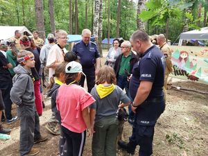 Zdjęcie w kolorze, przedstawia grupę  dzieci i młodzieży - uczestników obozu, obok stoi policyjny przewodnik psa służbowego, w tle namioty harcerskie.