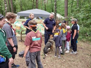 Zdjęcie w kolorze, przedstawia grupę  dzieci i młodzieży - uczestników obozu razem z policyjnym przewodnikiem psa służbowego i policjantką, w tle namioty harcerskie.