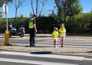 Zdjęcie w kolorze przedstawia przejście dla pieszych po ktorym dzieci po prawej ida w towarzystwie   policjanta po lewej. Wszyscy maja kamizelki odblaskowe.