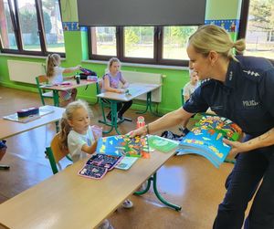Zdjęcie w kolorze przedstawia policjantkę wręczajacą książeczkę &quot; Bezpieczna droga do szkoły &quot; uczennicy  siedzącehjw szkolnej ławce.