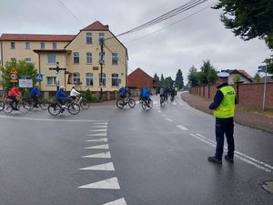 Zdjęcie w kolorze przedstawia policjanta stojacego na skrzyżowaniu, dalej przejeżdżający rowerzysci w tle zabudowania.