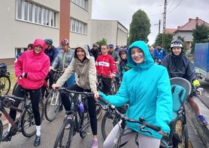 Zdjęcie w kolorze przedstawia na pierwszym planie kobiety w różnokolorowych kurtkach przeciwdeszczowych stojące z rowerami , tuz za nimi pozostali uczestnicy rajdu.