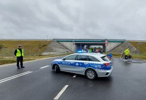 Zdjęcie w kolorze przedstawia Policjanta stojącego na DW 487 obok stoi radiowóz, za nimi jadący peleton.