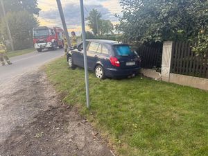 Zdjęcie w kolorze przedstawia granatowy samochód  zaparkowany przy ogrodzeniu. Pojazd w kadrze od tyłu nie widać uszkodzeń. W tle pojazd Straży Pożarnej.