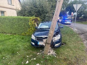 Zdjęcie w kolorze przedstawia widok od przodu - granatowy samochód zaparkowany przy ogrodzeniu oraz wkomponowany przednim zderzakiem w drewniany słup w betonowym  pokruszonym filarze. Samochód posiada uszkodzony przedni zderzak i pokrywę silnika. W tle policyjny samochód z włączonymi światłami błyskowymi.