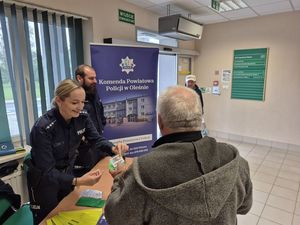 Zdjęcie w kolorze przedstawia wnętrze ZUS para policjantów wręcza element odblaskowy mężczyźnie w szarej kurtce, w tle plakat rozwijany z rolki z napisem Komenda Powiatowa Policji w Oleśnie.