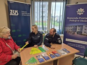 Zdjęcie w kolorze przedstawia wnętrze ZUS para policjantów wręcza element odblaskowy kobiecie  w czerwonej kurtce, w tle dwa plakaty rozwijane z rolki z lewej  napisem  ZUS i po prawej z napisem Komenda Powiatowa Policji w Oleśnie.