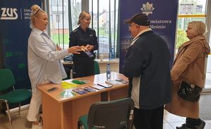 Zdjęcie w kolorze przedstawia wnętrze ZUS dwie kobiety w tym jedna policjantka  wręcza ulotkę informacyjną mężczyźnie w ciemnej kurtce, w tle plakaty rozwijane z rolki po lewej z napisem ZUS i po prawej z napisem Komenda Powiatowa Policji w Oleśnie.