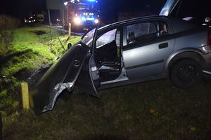 Zdjęcie w kolorze przedstawia rozbity pojazd osobowy w rowie, za nim na jezdni pojazd straży pożarnej z włączonymi sygnałami świetlnymi.