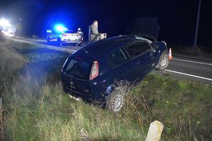 Zdjęcie w kolorze przedstawia rozbity pojazd częściowo na jezdni i na poboczu, dalej wóz policji i straży pożarnej z włączonymi sygnałami świetlnymi.