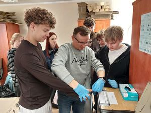 Zdjęcie w kolorze przedstawia młodzież podczas zajęć w pracowni technika kryminalistyki. Policjant w ubraniu cywilnym demonstruje jak prawidłowo wykonać odbitki palców.