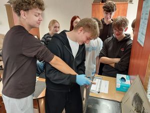 Zdjęcie w kolorze przedstawia młodzież podczas zajęć w pracowni technika kryminalistyki. Uczniowie sami wykonują odbitki palców.
