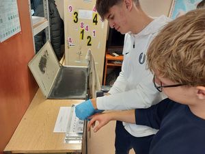 Zdjęcie w kolorze przedstawia młodzież podczas zajęć w pracowni technika kryminalistyki. Uczniowie sami wykonują odbitki palców.