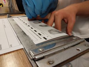 Zdjęcie w kolorze przedstawia młodzież podczas zajęć w pracowni technika kryminalistyki. Uczniowie sami wykonują odbitki palców.