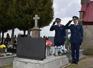 Zdjęcie w kolorze przedstawia dwóch policjantow w mundurach galowych, oddających honor - trzymających prawe dłonie przy czapkach. W tle mogiły krzewy i niebo.