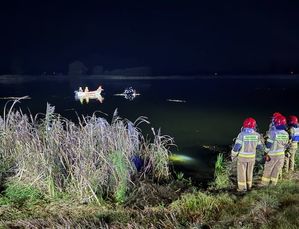 Zdjęcie w kolorze przedstawia stojących na brzegu strażaków w oddali zarys łódki ratowniczej oraz wystający fragment podtopionego w stawie pojazdu, do koła panuje zmrok.