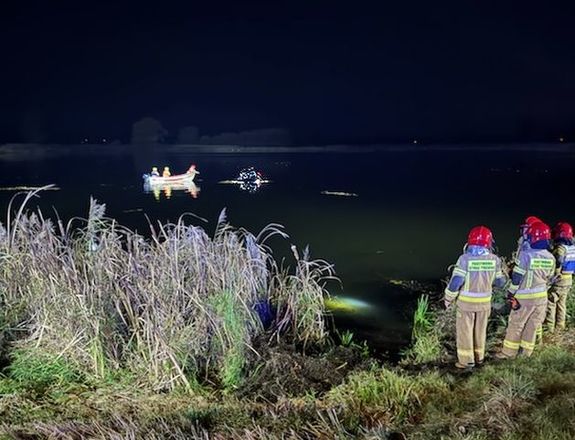 Zdjęcie w kolorze przedstawia stojących na brzegu strażaków w oddali zarys łódki ratowniczej oraz wystający fragment podtopionego w stawie pojazdu, do koła panuje zmrok.