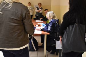 Zdjęcie w kolorze przedstawia na pierwszym planie pracowników cywilnych a za nimi przy stołach policjanci przy punkcie medycznym.