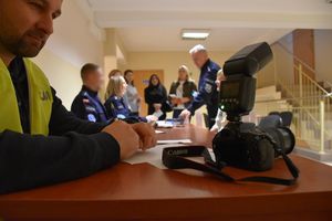 Zdjęcie w kolorze przedstawia policjanta- fotografa siedzącego przy stole na którym znajduje sie aparat fotograficzny.