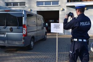 Zdjęcie w kolorze przedstawia stanowisko do przyjęcia pojazdów obsługiwane przez policjanta, który fotografuje pojazd typu bus.