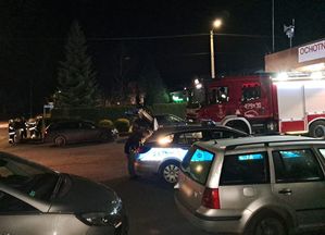 Zdjęcie w kolorze przedstawia pojazdy osobowe, za którymi po prawej wystaje fragment radiowozu, mężczyzna wkłada coś do bagażnika.