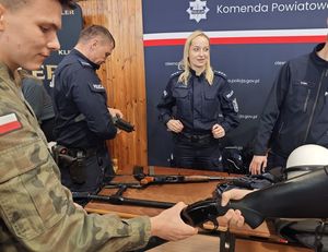 Zdjęcie w kolorze przedstawia stoisko policyjne z funkcjonariuszami, którzy promują służbę demonstrując uczniom jednostki broni i elementy wyposażenia.