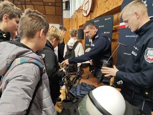 Zdjęcie w kolorze przedstawia stoisko policyjne z funkcjonariuszami, którzy promują służbę demonstrując uczniom jednostki broni i elementy wyposażenia.