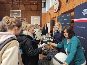 Zdjęcie w kolorze przedstawia stoisko policyjne z funkcjonariuszami oraz kobietę w ubraniu cywilnym, którzy promują służbę demonstrując uczniom jednostki broni i elementy wyposażenia.