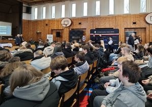 Zdjęcie w kolorze przedstawia salę gimnastyczną wypełnioną uczniami siedzącymi na krzesłach, w tle stoisko policyjne z funkcjonariuszami.