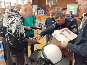 Zdjęcie w kolorze przedstawia stoisko policyjne z funkcjonariuszami, którzy promują służbę demonstrując uczniom jednostki broni i elementy wyposażenia.
