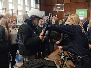 Zdjęcie w kolorze przedstawia stoisko policyjne z funkcjonariuszami, którzy promują służbę demonstrując uczniom jednostki broni i elementy wyposażenia.