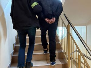 Zdjęcie w kolorze przedstawia, mężczyznę w kajdankach zapiętych na ręce trzymane z tyłu, który jest prowadzony za rękę przez policjanta. Obaj wchodzą po schodach.