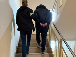 Zdjęcie w kolorze przedstawia dwóch mężczyzn idących po schodach , po prawej to zatrzymany ma założone kajdanki na ręce trzymane z tyłu, po lewej policjant ubrany po cywilnemu z opaską z napisem policja na prawym ramieniu.