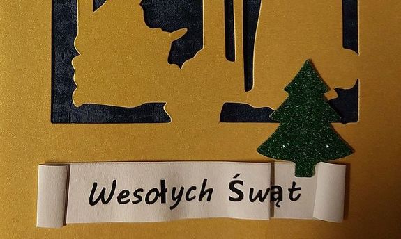 Zdjęcie w kolorze przedstawia kontury stajenki betlejemskiej, pod spodem naklejka zielona choinka i pod nią napis Wesołych Świąt.