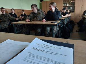 Zdjęcie w kolorze przedstawia kartki testu leżące na ławce w szkolnej klasie w tle uczniowie siedzący w ławkach.