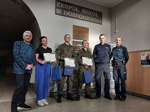 Zdjęcie w kolorze przedstawia uczniów klas mundurowych wspólnie z policjantami i dyrektorem pozujących do zdjęcia. Za nimi na ścianie napis Zespół Szkół w Dobrodzieniu.