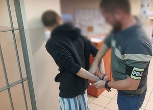 Zdjęcie w kolorze przedstawia policjanta , który stoi za mężczyzną który ma kajdanki założone na ręce trzymane z tyłu.