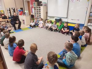 Zdjęcie w kolorze, przedstawia grupę przedszkolaków siedzących na podłodze w kole, po lewej stronie na środku znajduje się policjant.