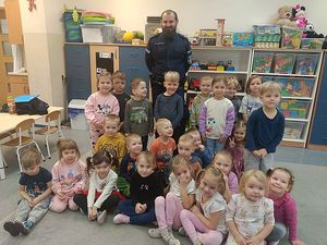 Zdjęcie w kolorze, przedstawia grupę przedszkolaków siedzących na podłodze po środku znajduje się policjant.