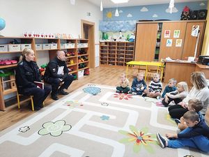 Zdjęcie w kolorze przedstawia grupę przedszkolaków siedzących na podłodze naprzeciw nich para policjantów.