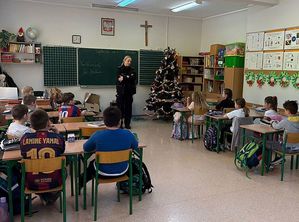 Zdjęcie w kolorze przedstawia, uczniów siedzących w klasie, na środku stoi policjantka.