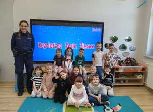 Zdjęcie w kolorze przedstawia dzieci siedzące na dywanie obok po lewej stoi policjantka za nimi ekran z napisem Bezpieczne Ferie.