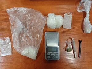 Zdjęcie w kolorze, przedstawia worki foliowe z białym proszkiem oraz mały woreczek foliowy z suszem roślinnym, obok fajka szklana do papierosów oraz waga jubilerska. Wszystkie przedmioty położone na drewnianym blacie.