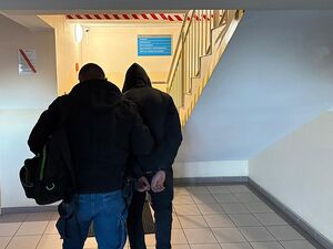 zdjęcie w kolorze przedstawia dwóch mężczyzn idących w stronę schodów, po lewej nieumundurowany policjant trzyma pod ramię mężczyznę po prawej który ma kajdanki zapięte na rękach trzymanych z tyłu.
