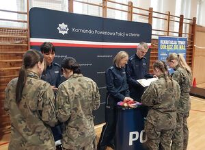 Zdjęcie w kolorze przedstawia młode kobiety w mundurach koloru zielonego - moro, stoją przy stoisku  promocyjnym z policjantkami.