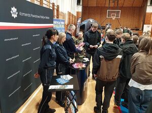 Zdjęcie w kolorze przedstawia uczniów szkół, stoją przy stoisku  promocyjnym z policjantkami, w tle sala gimnastyczna.
