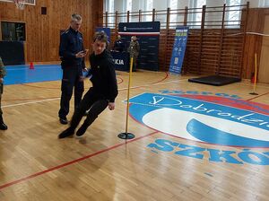 Zdjęcie w kolorze przedstawia ucznia podczas biegu po kopercie, za nim policjant, który mierzy czas.