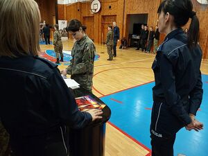 Zdjęcie w kolorze przedstawia na pierwszym planie policjantki, za nimi w tle uczniowie w mundurach wojskowych na sali gimnastycznej.