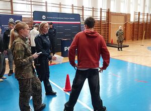 Zdjęcie w kolorze przedstawia uczniów szkół, stoją  z policjantkami, w tle sala gimnastyczna.